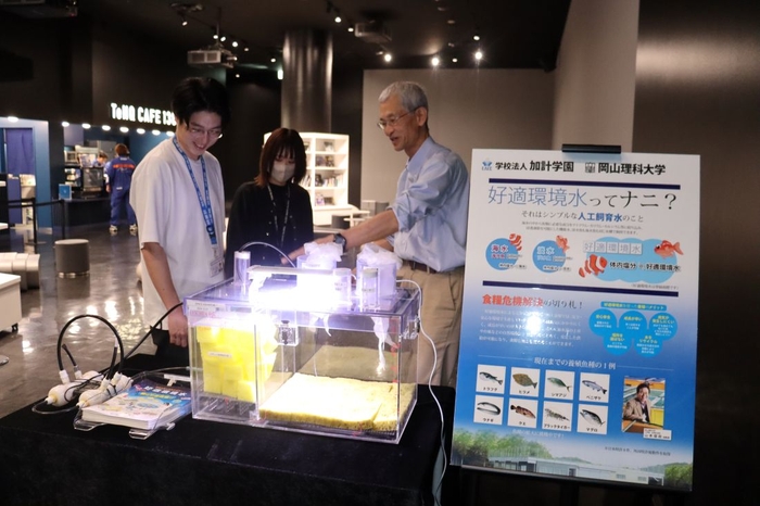 会場に水槽を設置してTeNQのスタッフと打ち合わせる津村誠一・生物生産教育研究センター招聘教授（右）＝5月20日