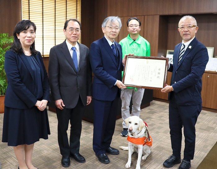 高野センター長（右端）から感謝状を受け取る平野学長（右から3人目）。2人の間で座っているのがPR犬・アクア