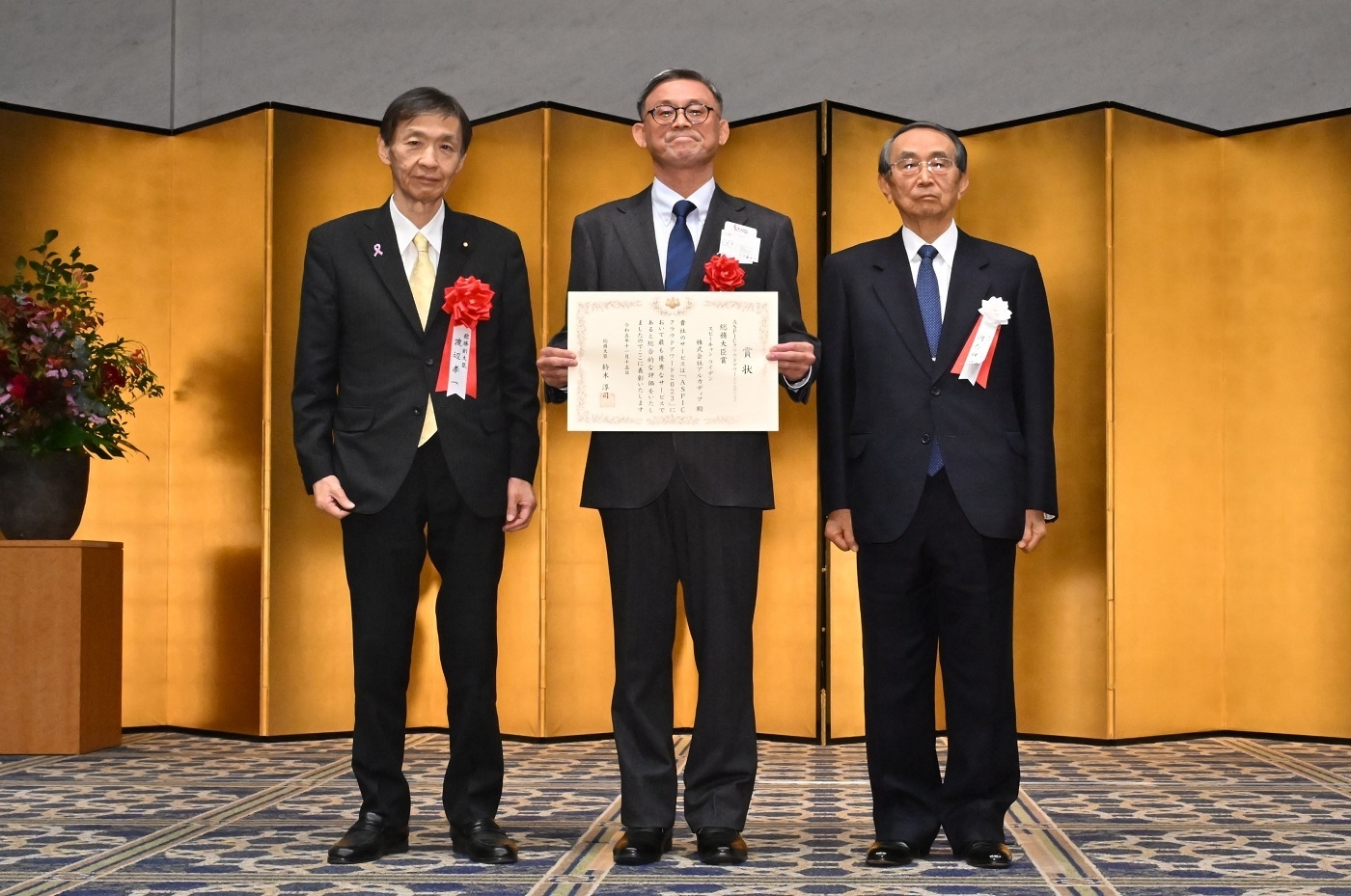 総務大臣賞授与(写真左:総務副大臣 渡辺孝一様 中央:株式会社アルカディア様 右:河合会長)